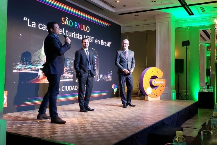 Secretário Luiz Alvaro fala ao microfone, em cima do palco, durante o evento. Ao lado dele estão o Secretário Adjunto de Turismo e o Presidente da Câmara LGBT.